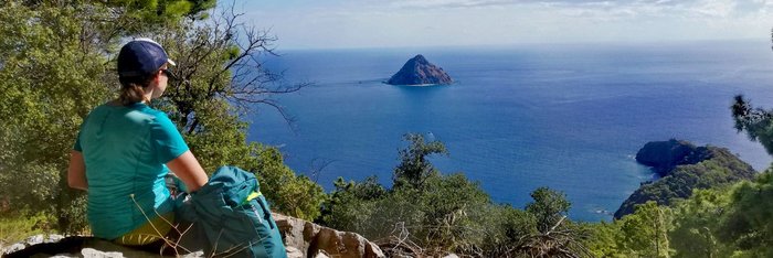 Eine Frau sitzt auf einem steinigen Weg und blickt auf einen Felsen mitten im offenen Meer